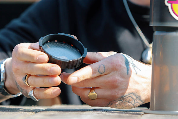 The image shows someone holding a metal filter for an AeroPress coffee maker. The AeroPress is a popular device for brewing coffee, and metal filters like the one in the picture can be used as an alternative to the standard paper filters. They are reusable and often favoured by those looking for a more environmentally friendly option or a slightly different flavour profile in their coffee. The metal filter allows more oils and fine particles to pass through, resulting in a fuller-bodied cup of coffee.